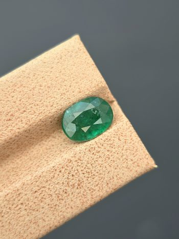 A well-lit, professional image of a 1.68 carat natural emerald from the Swat Valley, Pakistan. The gemstone has an oval cut, a green color, and visible inclusions.